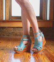 A woman wearing blue Princess wedge sandals with woven strappy uppers standing on a sustainable wooden floor.