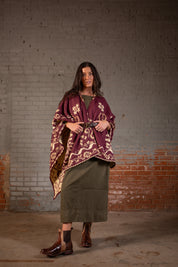 A woman stands before a gray and red brick wall wearing the Bed Stu Proem maroon patterned poncho, an olive green dress, brown boots, and a rustic casual belt.