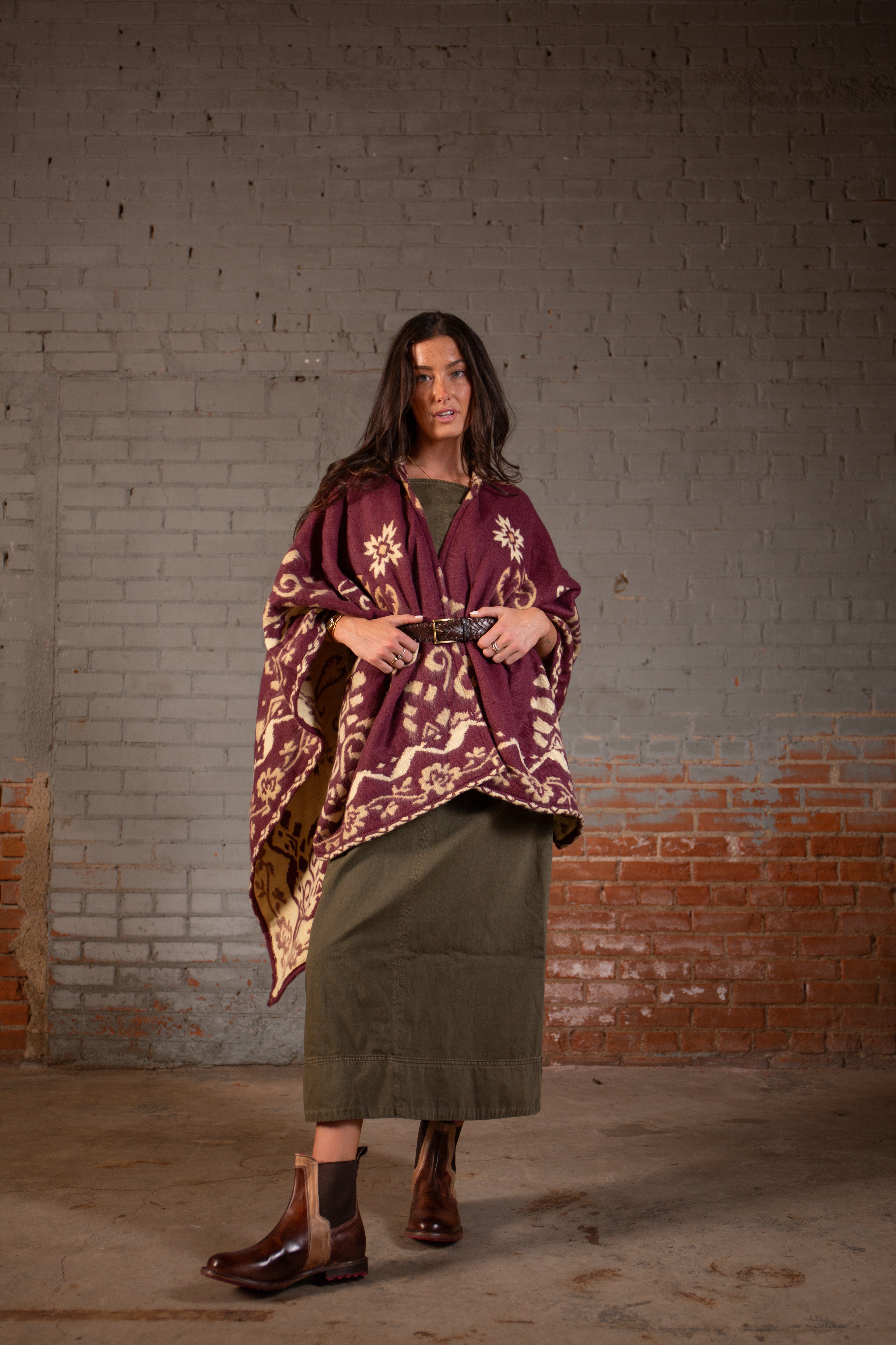 A woman stands before a gray and red brick wall wearing the Bed Stu Proem maroon patterned poncho, an olive green dress, brown boots, and a rustic casual belt.