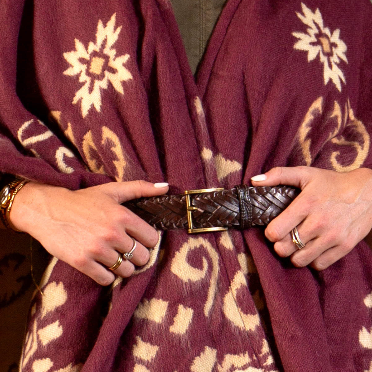 A person wearing the Bed Stu Proem rustic casual belt with a vintage metal buckle over a maroon patterned shawl, hands adorned with rings and manicured nails.