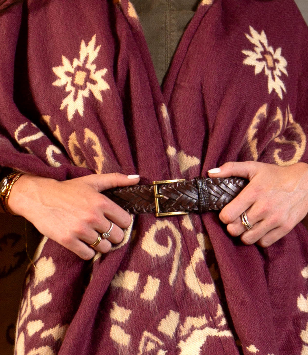A person wearing the Bed Stu Proem rustic casual belt with a vintage metal buckle over a maroon patterned shawl, hands adorned with rings and manicured nails.