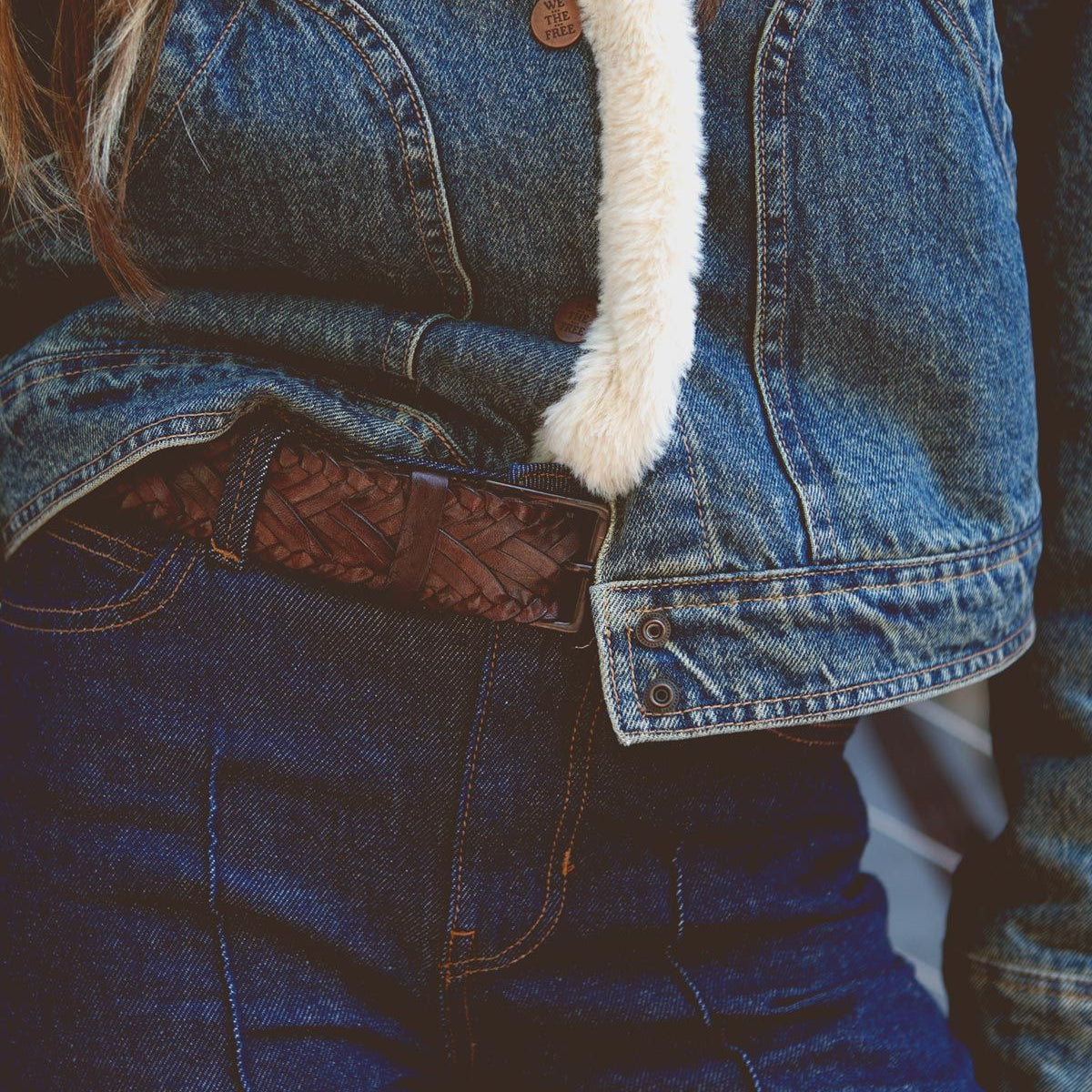 Close-up of a person wearing a denim jacket with a fuzzy lining and high-waisted jeans, cinched with the Proem belt by Bed Stu, featuring a dark brown texture and vintage metal buckle for that casual cool style.