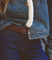 Close-up of a person wearing a denim jacket with a fuzzy lining and high-waisted jeans, cinched with the Proem belt by Bed Stu, featuring a dark brown texture and vintage metal buckle for that casual cool style.