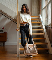A person in a cream sweater and black jeans stands by a staircase, holding a large metallic Bed|Stü Proper tote with handwoven straps.