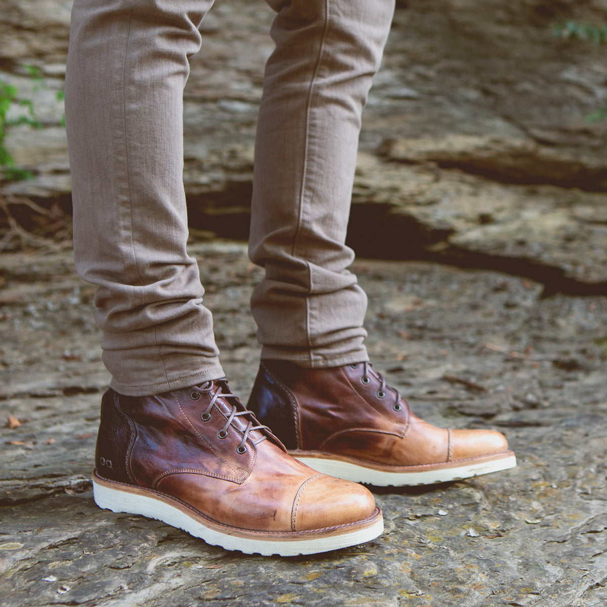 A person wearing Bed Stu men's leather boots and beige pants standing on a rocky surface.