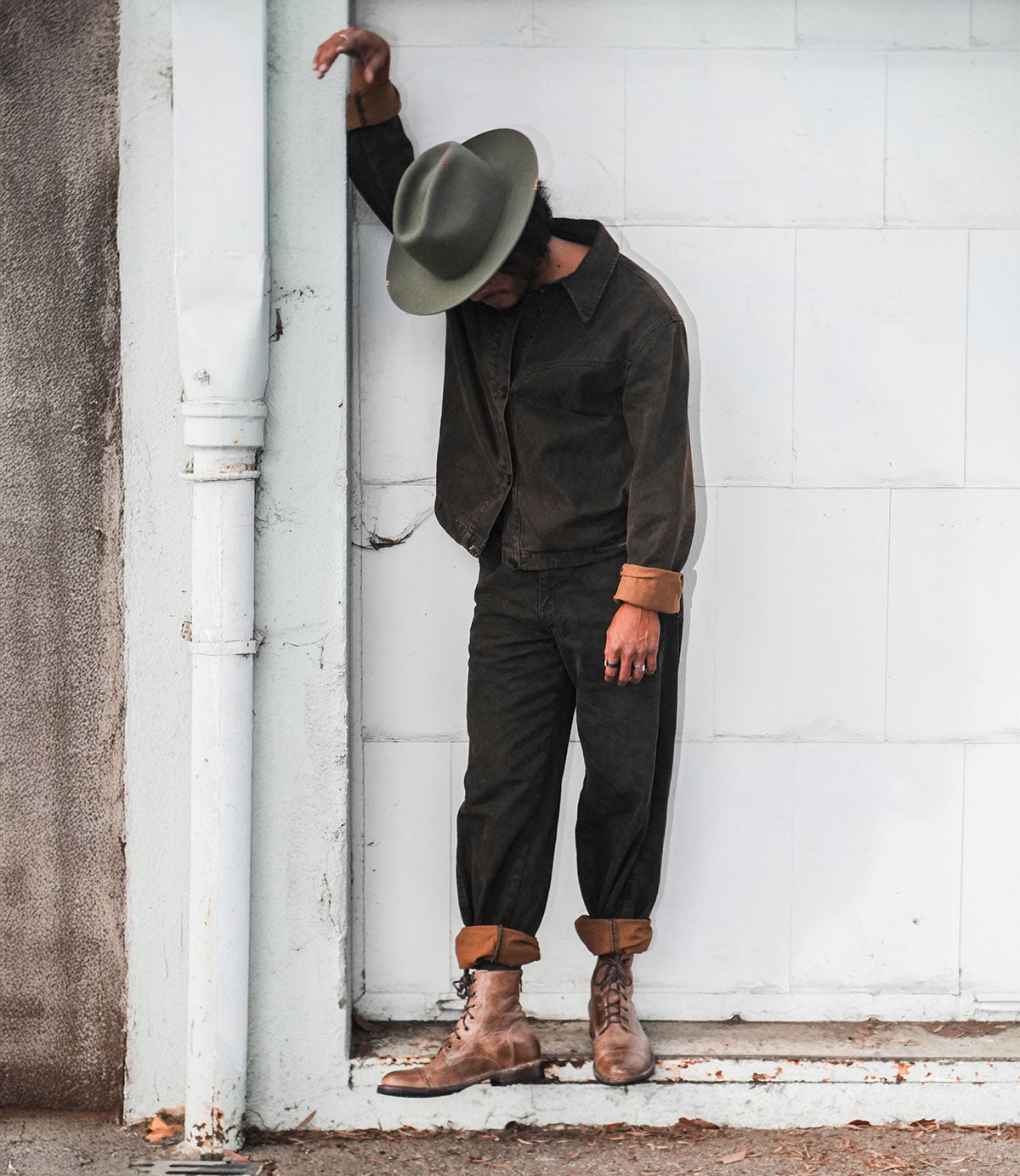 A person dressed in dark clothing and a hat leans against a white wall, looking downward, their Bed Stu Protege combat boots with Goodyear welt construction adding an edgy touch.