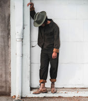 A person dressed in dark clothing and a hat leans against a white wall, looking downward, their Bed Stu Protege combat boots with Goodyear welt construction adding an edgy touch.