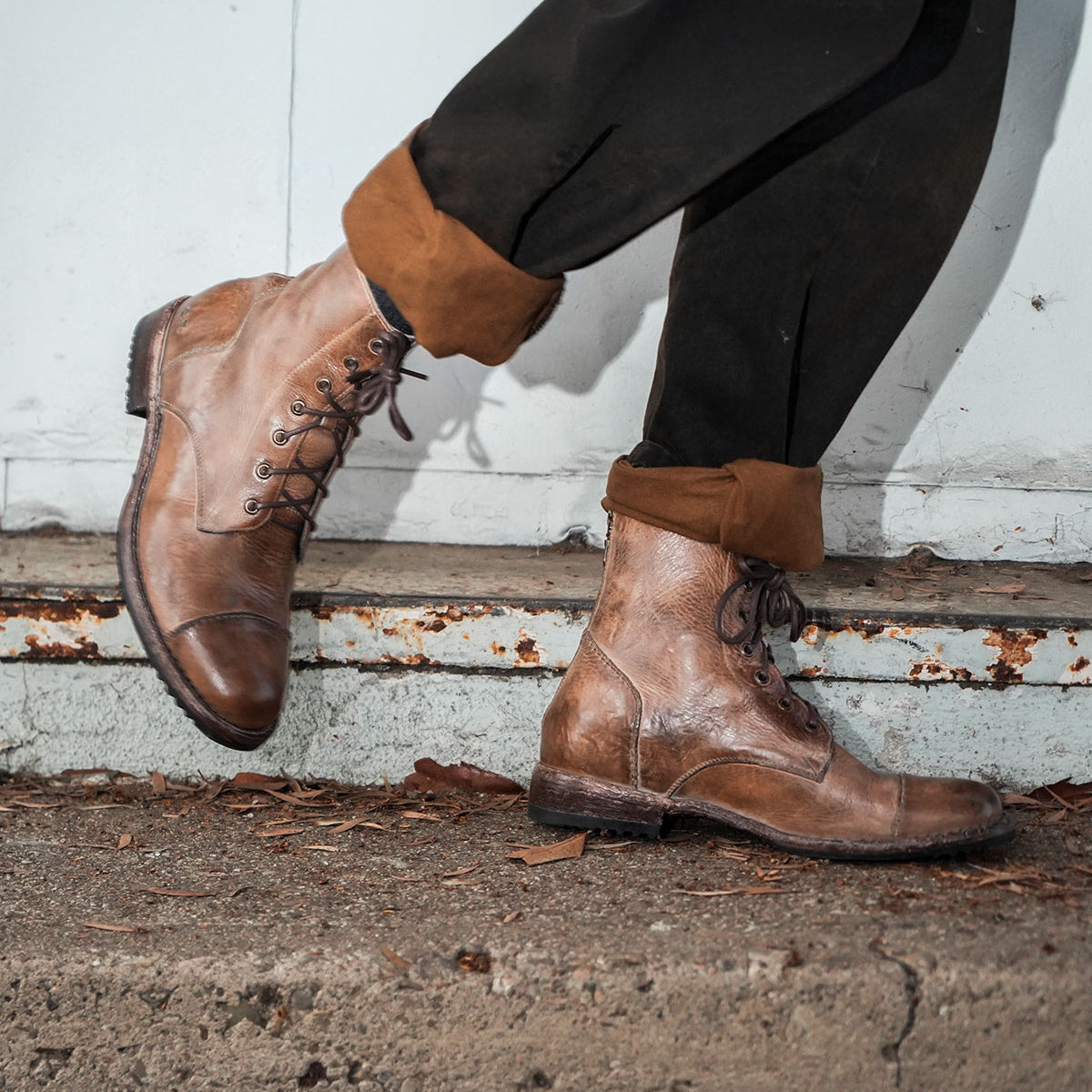 A person walks on a concrete surface near a metal wall wearing the Protege combat boots by Bed Stu, featuring a back zipper in brown, along with black pants.