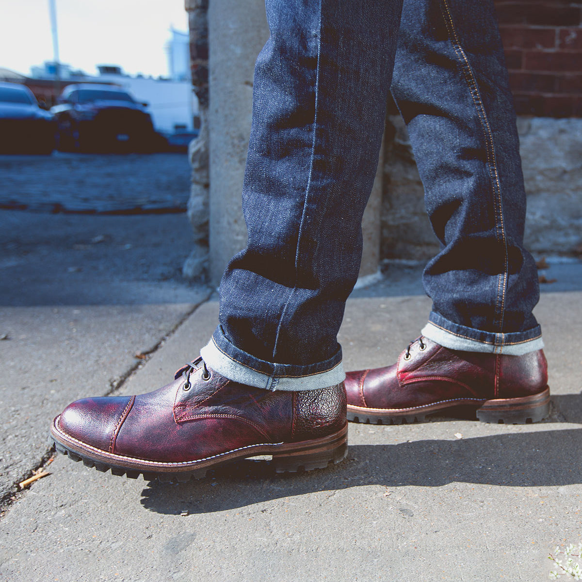 A person wearing dark jeans and Bed Stu's Protege Trek boots, featuring a Vibram outsole, stands confidently on a sidewalk.