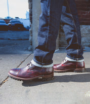 A person wearing dark jeans and Bed Stu's Protege Trek boots, featuring a Vibram outsole, stands confidently on a sidewalk.