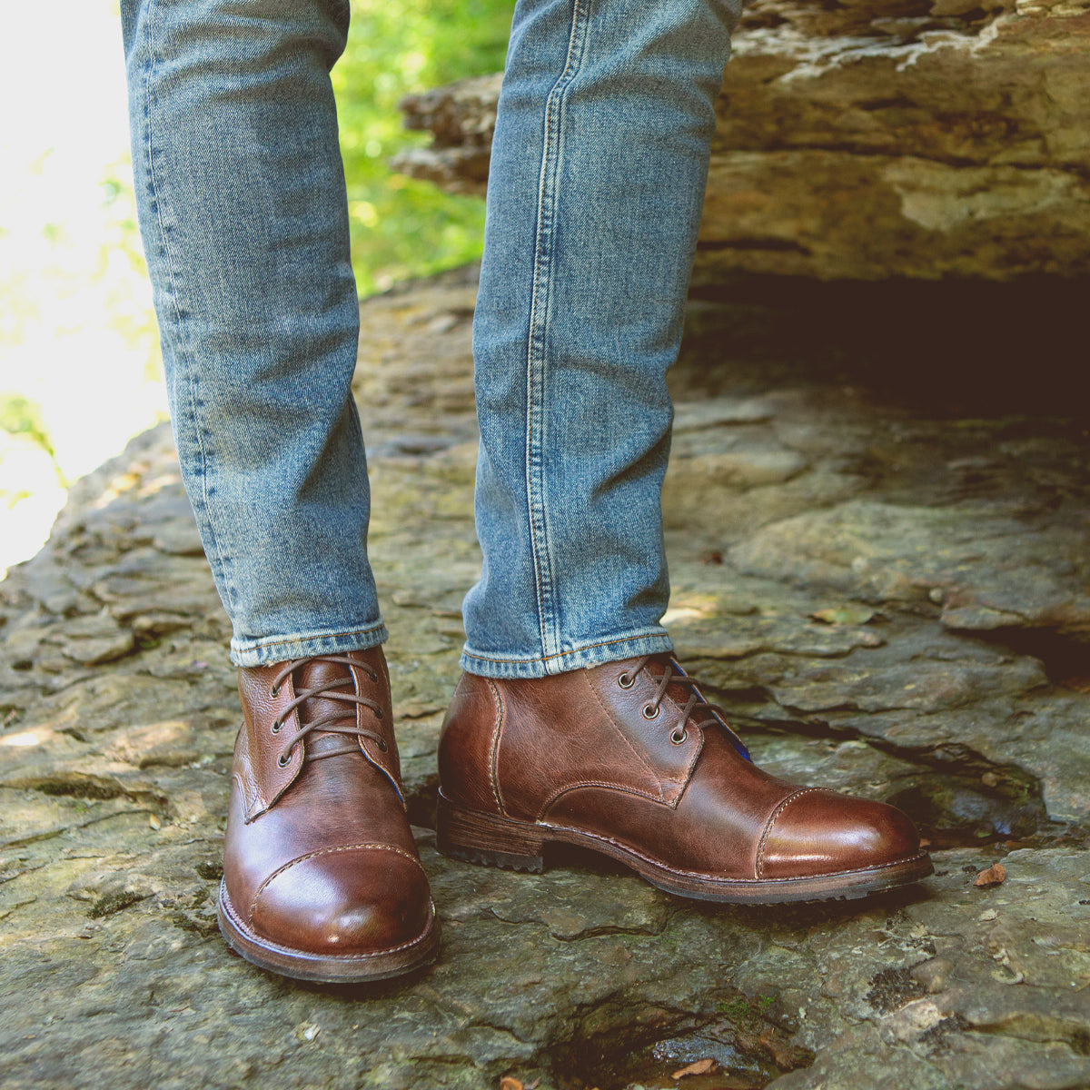 Person wearing rugged brown Protege boots from Bed Stu with Goodyear welt construction and blue jeans standing on a rocky surface.