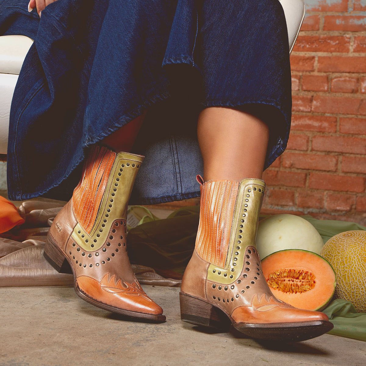 A woman wearing Bed Stu's Raise snip toe Western boots with dazzling studded accents and a denim skirt sits on a white chair. Nearby, two cantaloupes and a honeydew melon add to the rustic charm against a brick wall.