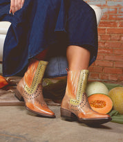A woman wearing Bed Stu's Raise snip toe Western boots with dazzling studded accents and a denim skirt sits on a white chair. Nearby, two cantaloupes and a honeydew melon add to the rustic charm against a brick wall.