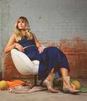 A person in a denim dress sits on a white chair against a brick wall, donning Bed Stu's Raise brown Western boots with elegant snip toes. The scene is enriched by fabric strewn around and two melons resting on the floor.