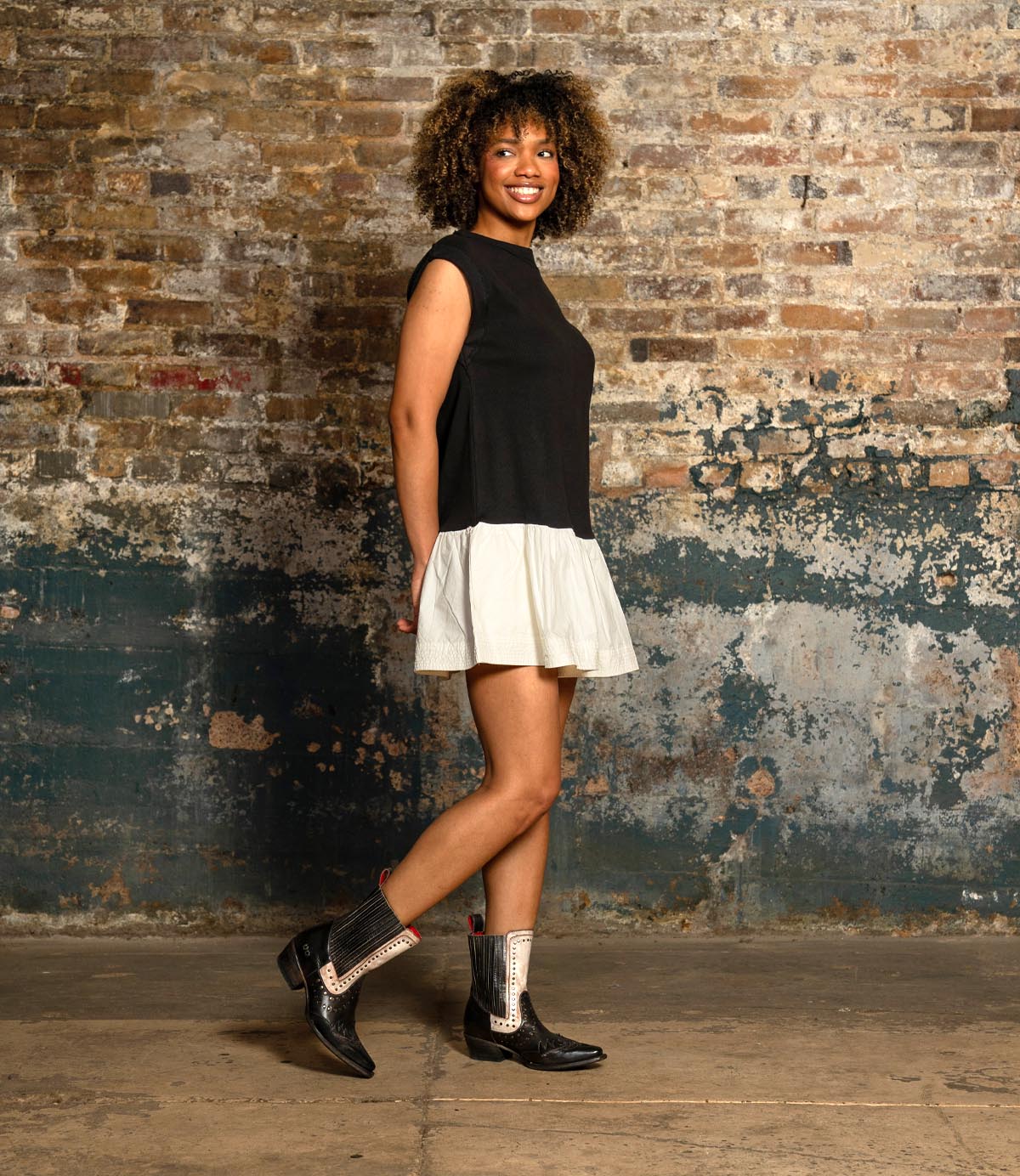 A woman stands sideways against a brick wall, smiling at the camera and showcasing her modern style in the Bed Stu "Raise" boots—sleek Western-inspired footwear in black and white.