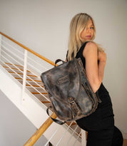 A woman with long blonde hair stands on the stairs, dressed in black and carrying the Bed|Stü Ranger backpack—crafted from sturdy brown leather—on her shoulder as she glances back toward the camera.