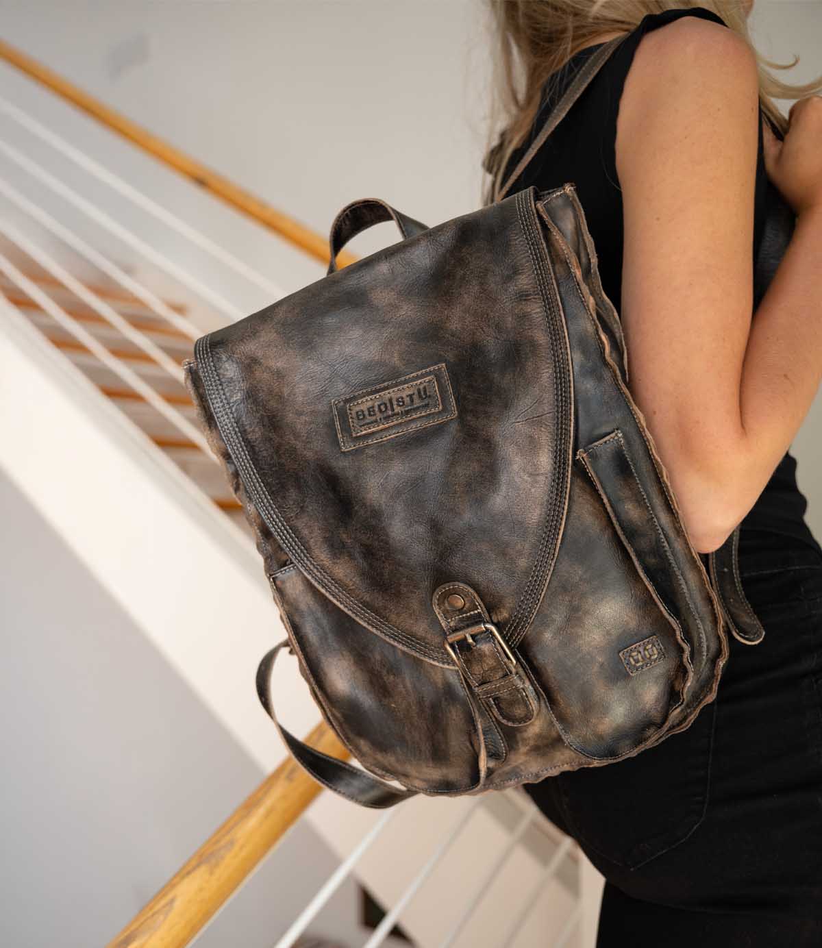 A person in black clothing stands on stairs, carrying the Ranger backpack by Bed|Stü—a sturdy, worn brown leather bag with a visible logo on the flap—making it an ideal choice for everyday wear.