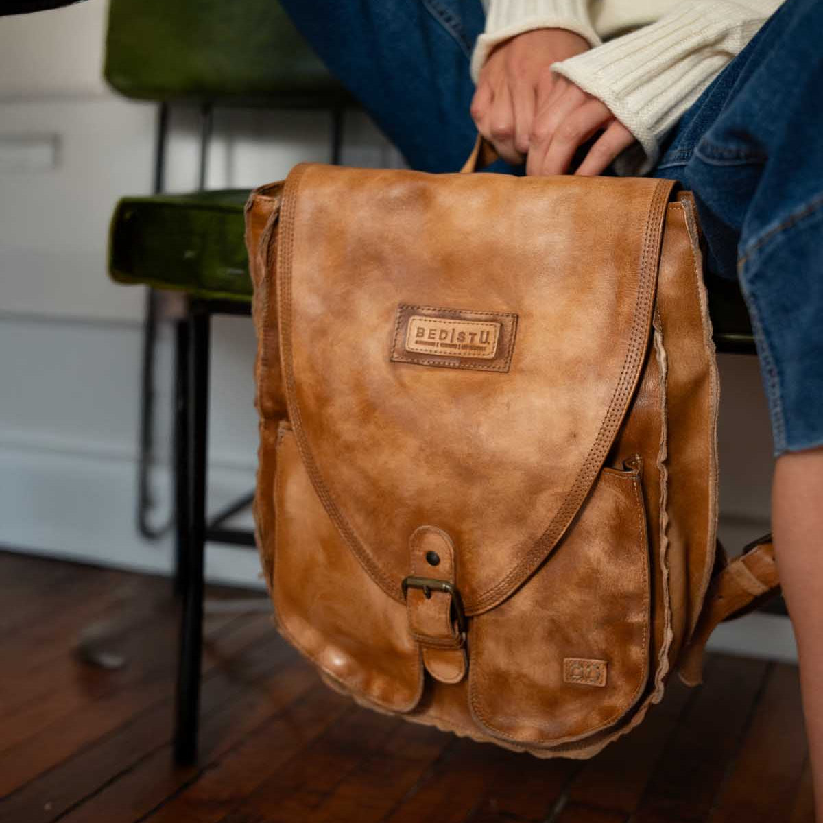 A person in jeans and a cream sweater sits on a chair, holding the Ranger brown leather backpack from Bed|Stü—perfect for everyday use—resting on a wooden floor.