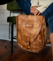 A person in jeans and a cream sweater sits on a chair, holding the Ranger brown leather backpack from Bed|Stü—perfect for everyday use—resting on a wooden floor.