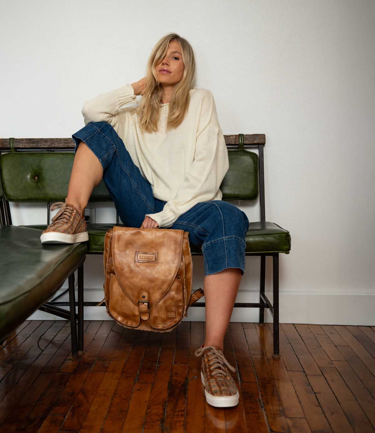A woman sits on a green bench, wearing a cream sweater, cropped jeans, and sneakers—perfect for everyday wear—with one leg up and the Bed|Stü Ranger sturdy brown leather backpack resting on the floor beside her.