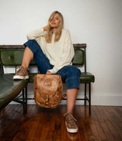 A woman sits on a green bench, wearing a cream sweater, cropped jeans, and sneakers—perfect for everyday wear—with one leg up and the Bed|Stü Ranger sturdy brown leather backpack resting on the floor beside her.