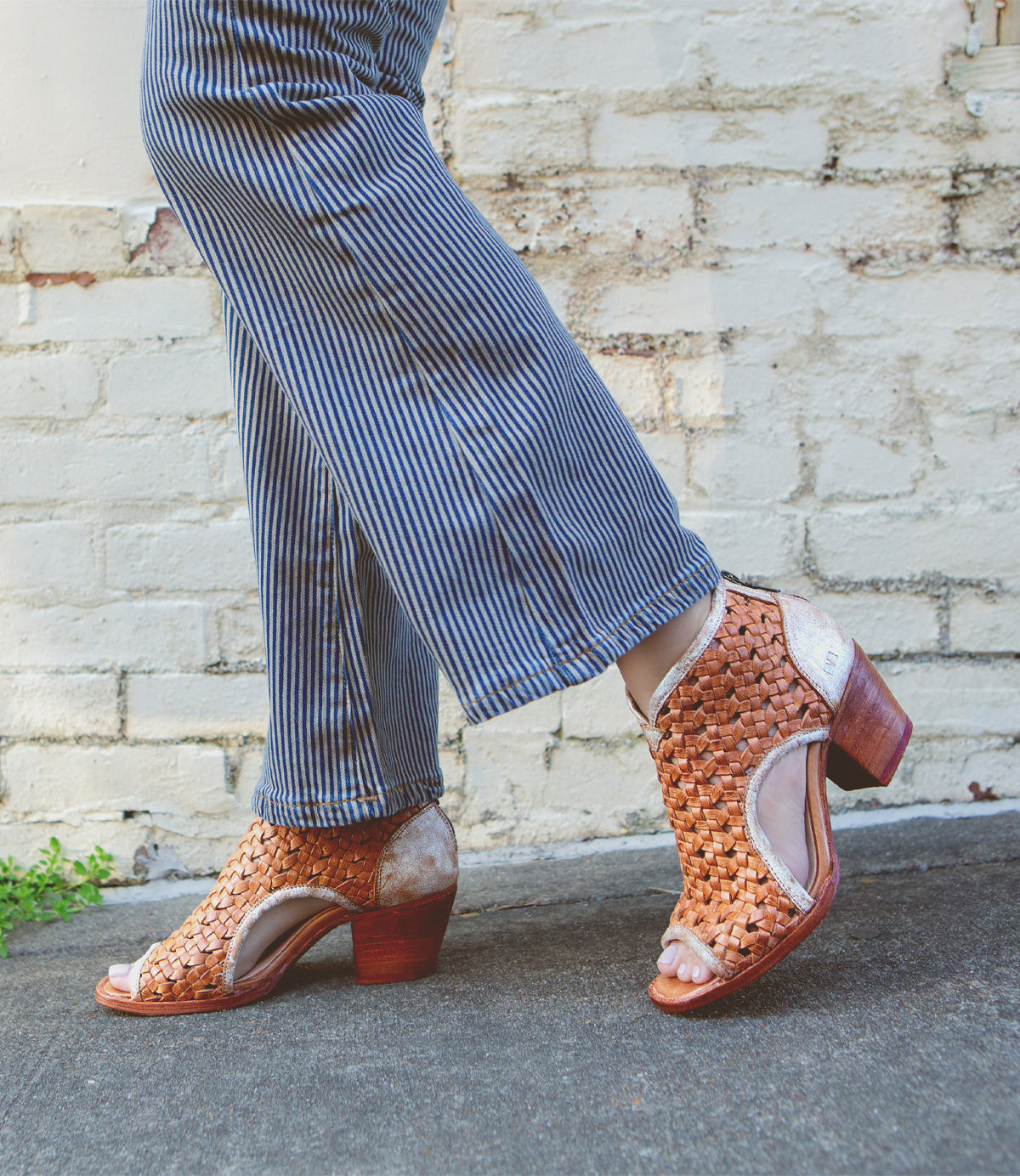 A person walking in stylish brown woven peep-toe heels crafted with vegetable tanned leather from Bed Stu, paired with striped trousers.