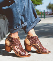 A person wearing the sophisticated, elegant Ransom shoes by Bed Stu—featuring brown woven open-toe heeled sandals—paired with rolled-up blue jeans, sits outside on a sunny day.