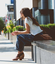 A woman in a white shirt and jeans sits on a city street bench, holding a small object in her hand. She wears handmade peep-toe heels and has the "Ransom" brown vegetable tanned leather bag by Bed Stu over her shoulder.