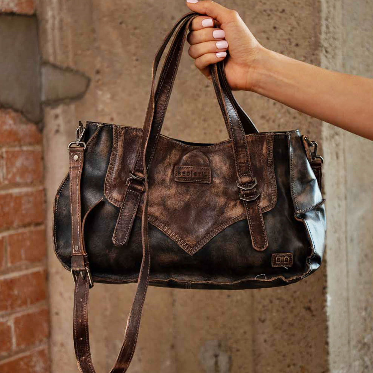 A hand holds the Bed|Stü Redrock handbag, crafted from distressed brown leather with double handles, a buckled handle, shoulder strap, and visible Bed|Stü branding, set against a concrete and brick background. This heritage-inspired design makes it the perfect compact bag for everyday use.