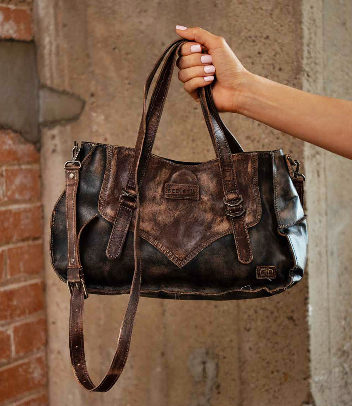 A hand holds the Bed|Stü Redrock handbag, crafted from distressed brown leather with double handles, a buckled handle, shoulder strap, and visible Bed|Stü branding, set against a concrete and brick background. This heritage-inspired design makes it the perfect compact bag for everyday use.