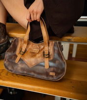 A person sits on wooden stairs holding the Bed|Stü Redrock, a brown leather handbag with two handles, highlighted by a buckled strap and visible brand accents. This compact everyday bag showcases a heritage-inspired design, effortlessly fusing classic style with modern functionality.