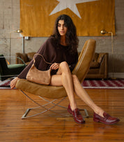 A woman sits in a chair holding a tan leather Reina shoulder bag, wearing burgundy leather penny loafers with a Goodyear welted construction.
