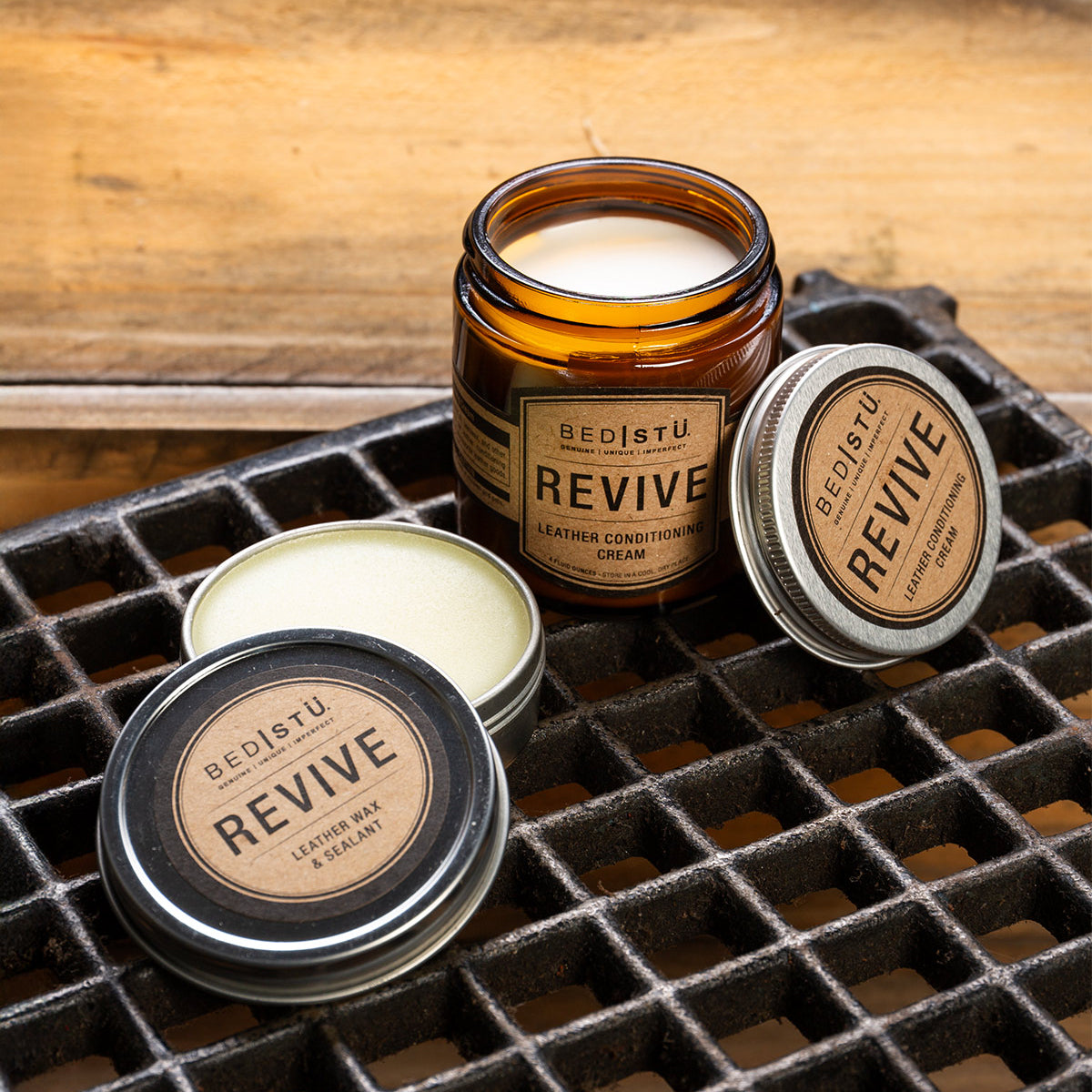 A jar and two tins of the Bed|Stü Revive Leather Care Package are displayed on a metal grid surface with a wooden background.