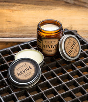 A jar and two tins of the Bed|Stü Revive Leather Care Package are displayed on a metal grid surface with a wooden background.