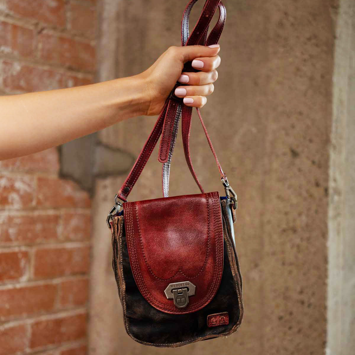 A hand holds up the Bed|Stü Rhyme, a compact crossbody bag crafted in two-tone brown and red leather with a metal clasp, against a brick and concrete background—offering a stylish hands-free accessory for any occasion.