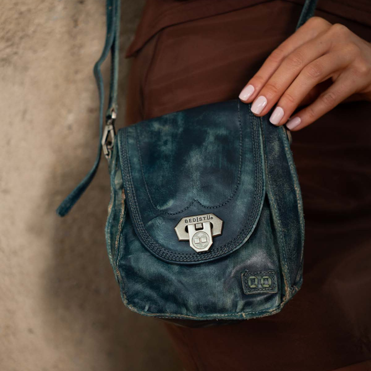 A person with manicured nails holds the “Rhyme” compact crossbody bag by Bed|Stü—crafted from weathered blue leather with a metal clasp—while standing against a textured wall.