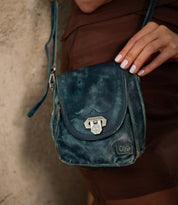 A person with manicured nails holds the “Rhyme” compact crossbody bag by Bed|Stü—crafted from weathered blue leather with a metal clasp—while standing against a textured wall.