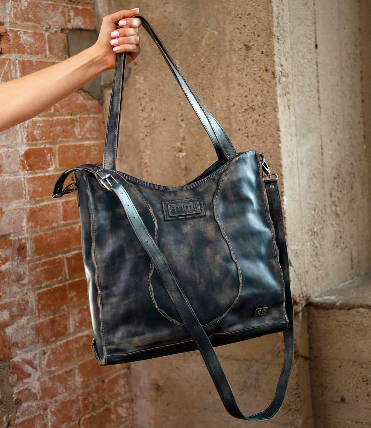 A hand holds the Bed|Stü Ridgefield tote, crafted from large, distressed black full-grain leather with two sturdy straps and a subtle embossed logo, set against brick and concrete walls. The stylish Ridgefield features a thoughtfully organized interior to keep daily essentials neatly in place.
