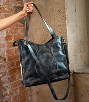 A hand holds the Bed|Stü Ridgefield tote, crafted from large, distressed black full-grain leather with two sturdy straps and a subtle embossed logo, set against brick and concrete walls. The stylish Ridgefield features a thoughtfully organized interior to keep daily essentials neatly in place.