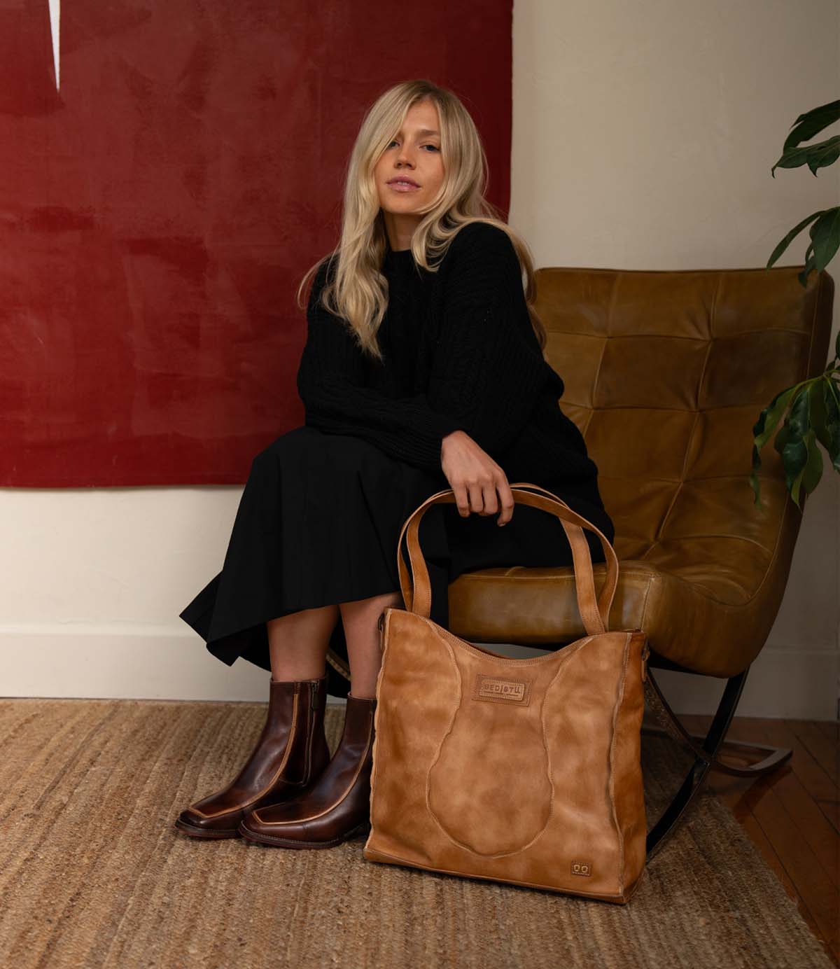 A woman in a black outfit sits on a brown leather chair, holding the Ridgefield daily tote by Bed|Stü, crafted from full-grain leather, with a red artwork and a plant in the background.