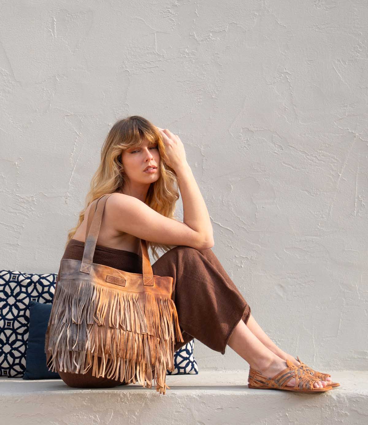 A woman with long hair sits on a ledge with patterned cushions, wearing a brown outfit and sandals, carrying the chic Bed Stu Rimes fringe tote against a light textured wall.