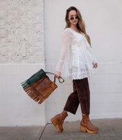A woman in a sheer white blouse, brown pants, and platform boots holds the Bed Stu Rimes handcrafted leather tote with green and fringe details, standing against a white wall.