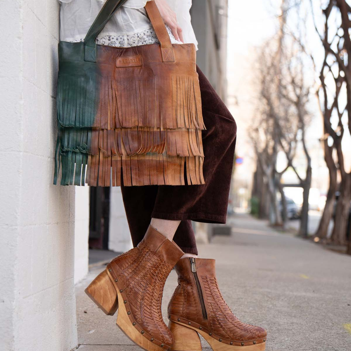 Wearing Bed Stu Rimes brown textured platform boots and cropped dark pants, a person holds a handcrafted leather tote with fringe detail and roomy interior while standing on a city sidewalk.
