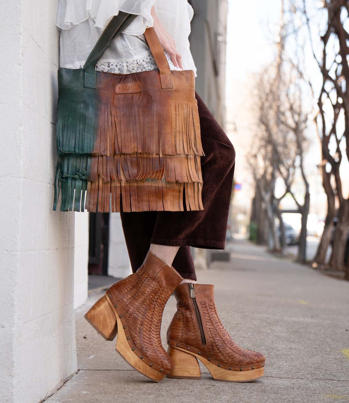 Wearing Bed Stu Rimes brown textured platform boots and cropped dark pants, a person holds a handcrafted leather tote with fringe detail and roomy interior while standing on a city sidewalk.
