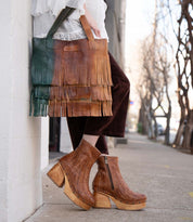 Wearing Bed Stu Rimes brown textured platform boots and cropped dark pants, a person holds a handcrafted leather tote with fringe detail and roomy interior while standing on a city sidewalk.
