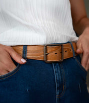 A person wearing a white textured top, blue jeans, and the Bed|Stü Riven heritage leather belt with a rectangular buckle, with hands in pockets.
