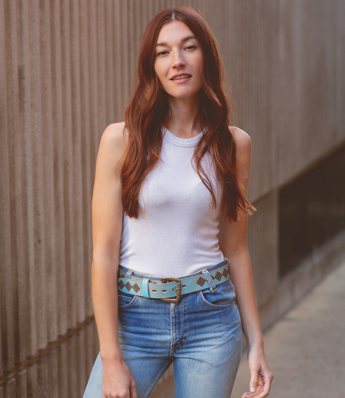 A woman with long red hair stands against a textured wall, wearing a white tank top, blue jeans, and a sophisticated Aztec belt from Bed Stu called Roam.