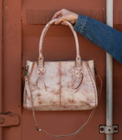 A hand elegantly holds the Rockababy by Bed Stu, a stylish everyday bag made from light brown leather with decorative stitching and laces, against a rust-colored metal backdrop. The person in a blue denim jacket perfectly blends comfort and style.