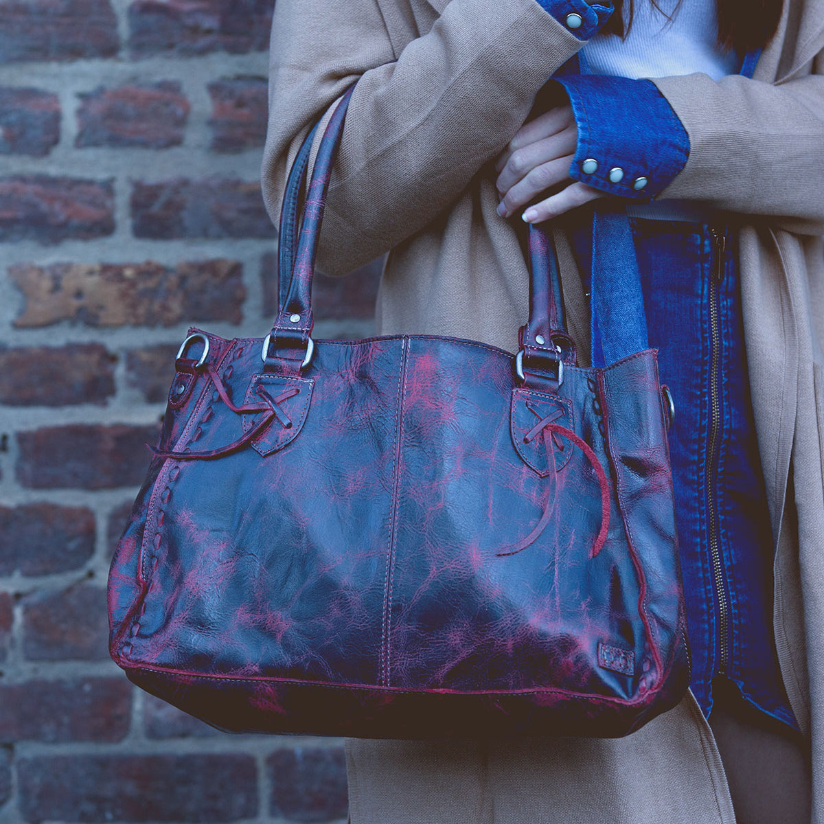 A person holding the Rockababy – a red leather everyday handbag with dual handles by Bed Stu, is wearing a tan coat and denim shirt while standing against a brick wall.