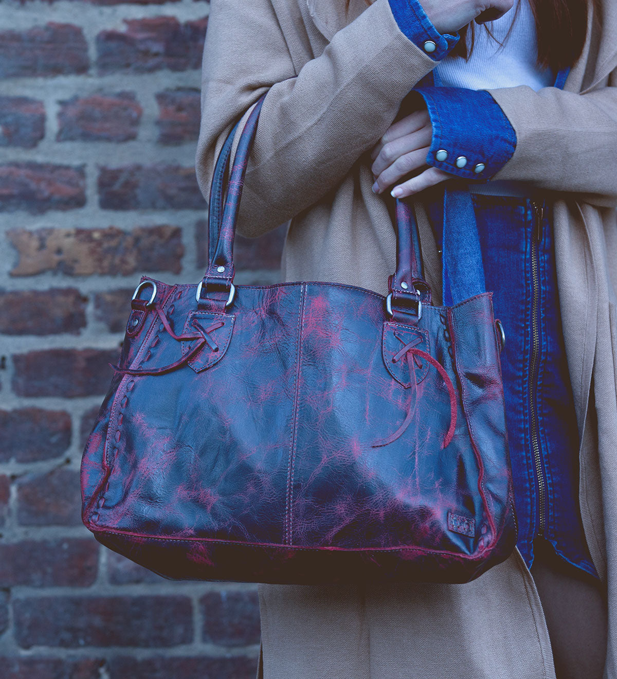 A person holding the Rockababy – a red leather everyday handbag with dual handles by Bed Stu, is wearing a tan coat and denim shirt while standing against a brick wall.